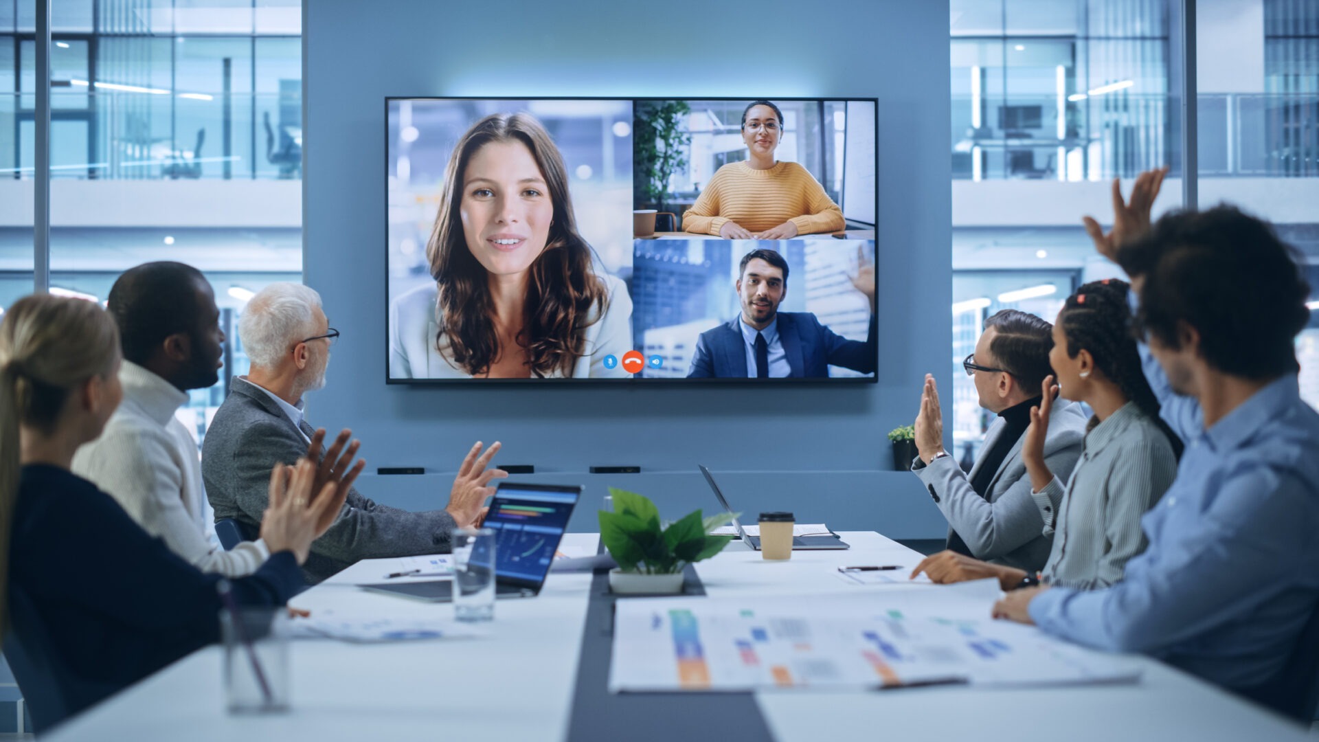 People in a modern conference room engage in a video call, displaying three persons on a large screen. Collaborative business setting.