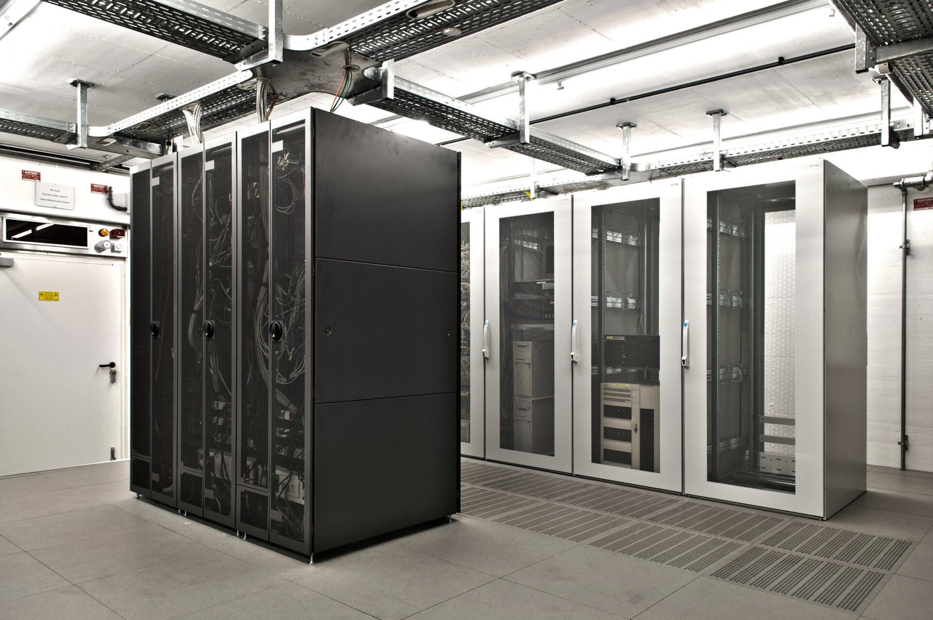 A modern server room features multiple black and white server racks lined up in a well-lit, industrial space with overhead cables.