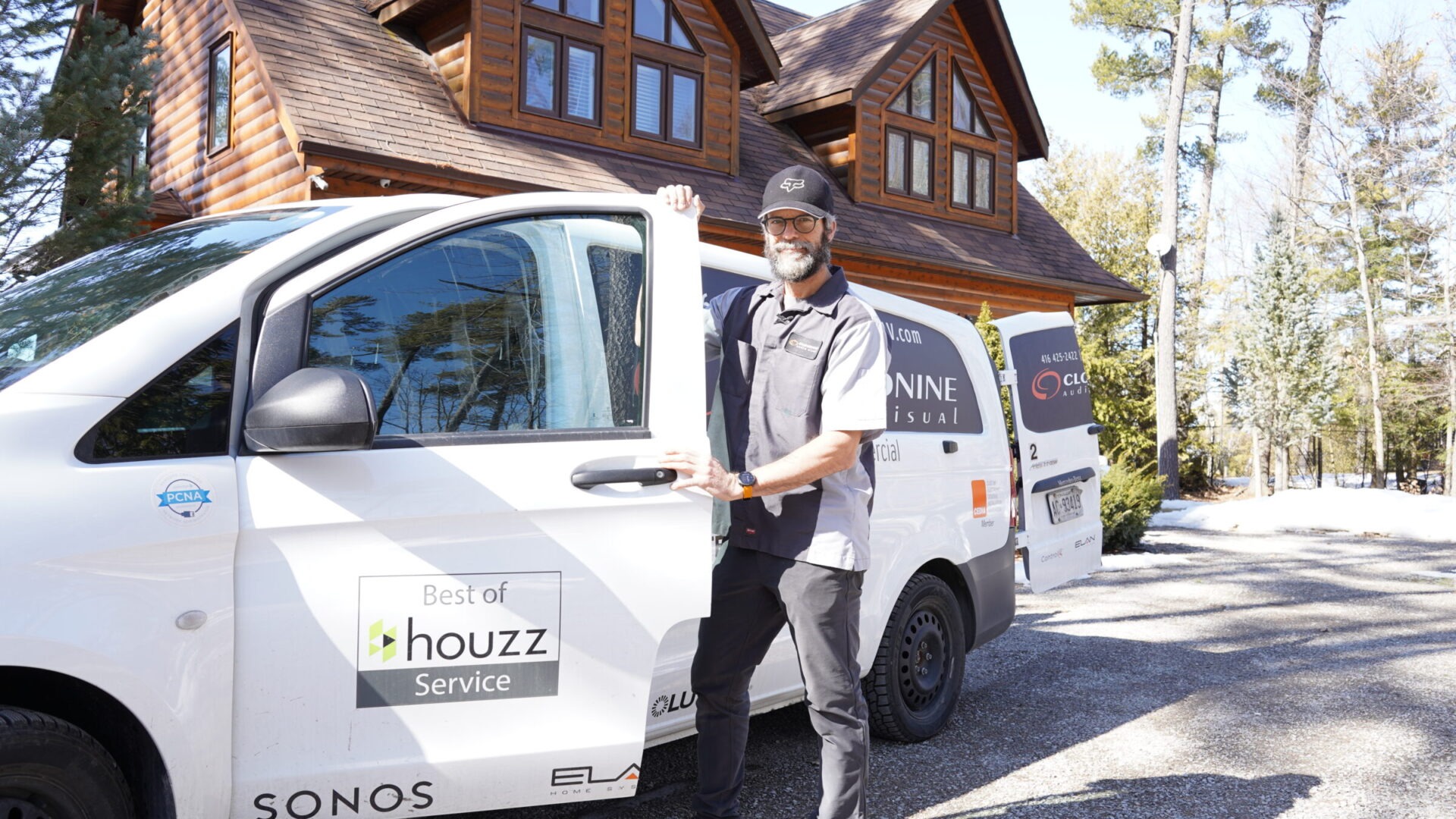 A person stands by a van labeled "houzz Service" in front of a wooden house, surrounded by trees on a sunny day.