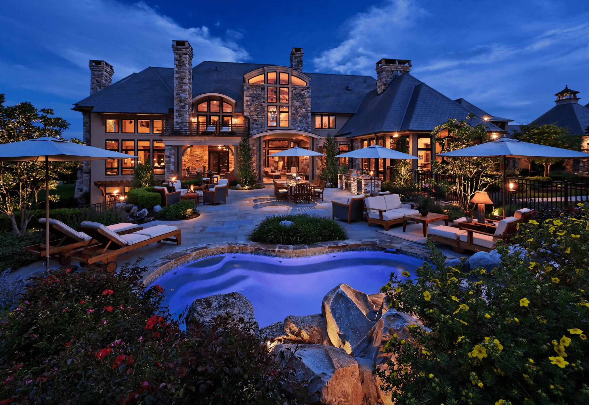 Luxurious estate at dusk with elegant stone architecture, illuminated pool, patio seating, and lush garden. Evening lights create a tranquil, inviting ambiance.
