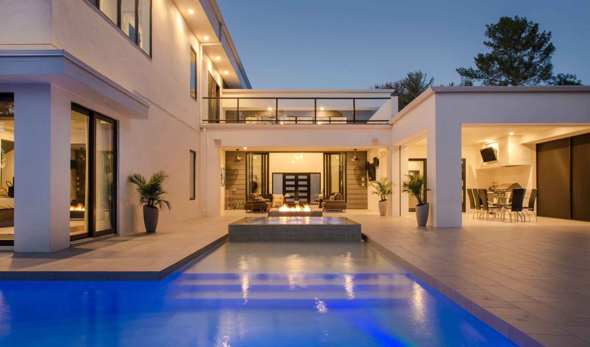 Modern luxury home with a pool, expansive patio, and stylish outdoor seating area at dusk, surrounded by trees and ambient lighting.