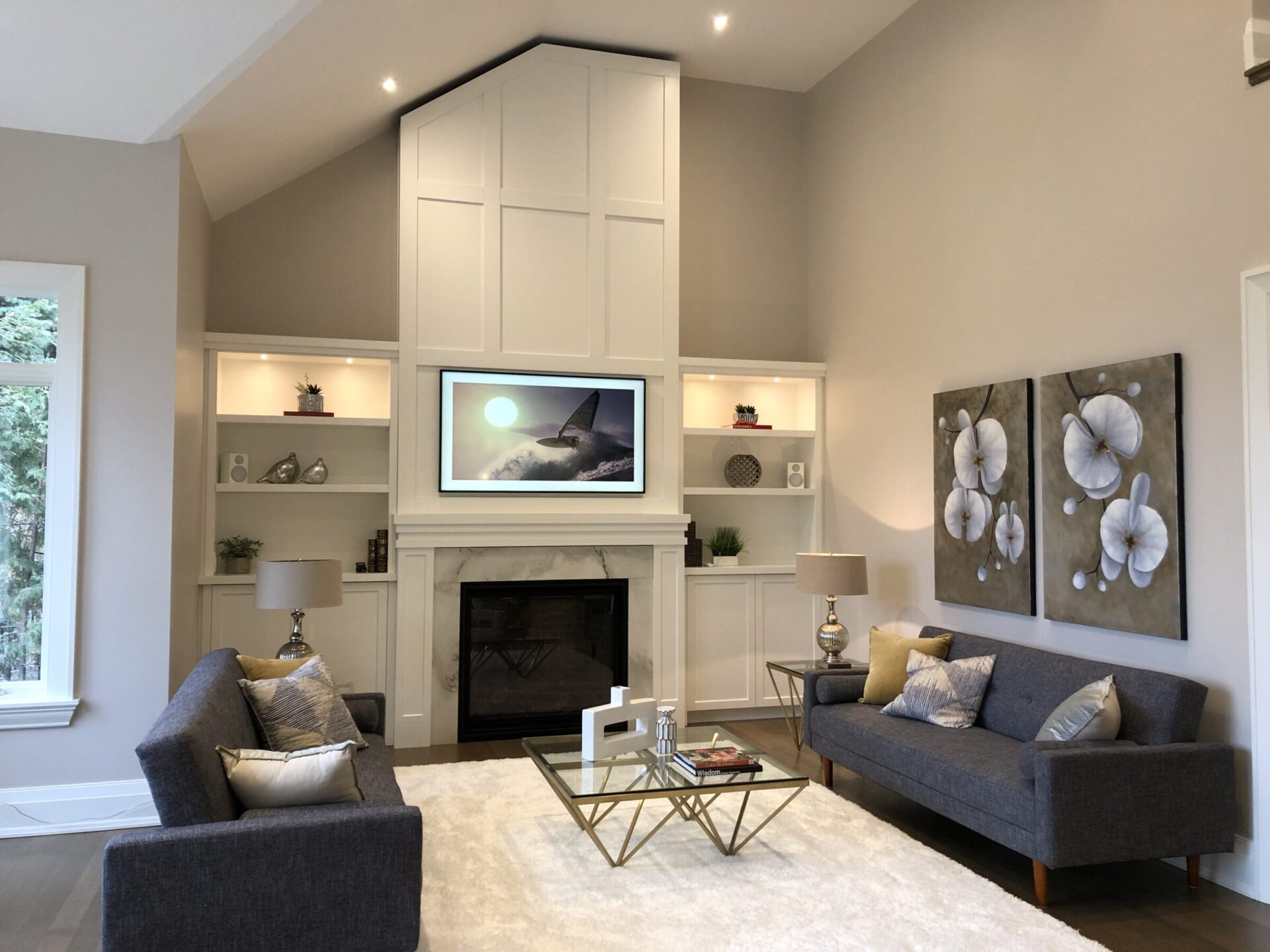 Modern living room with high ceiling, white walls, gray sofas, glass coffee table, and decorative fireplace. Large artwork and natural light enhance space.