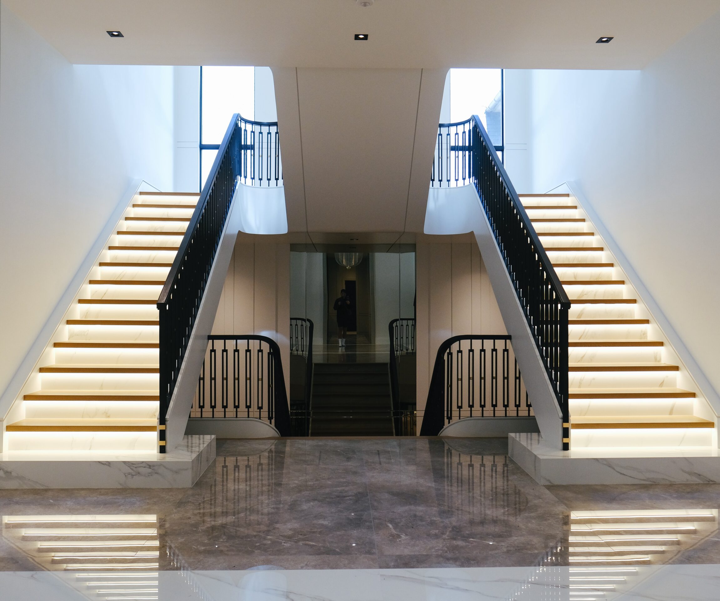 A modern interior with a split staircase, sleek design, reflective flooring, and a railing. A person stands at the top center.