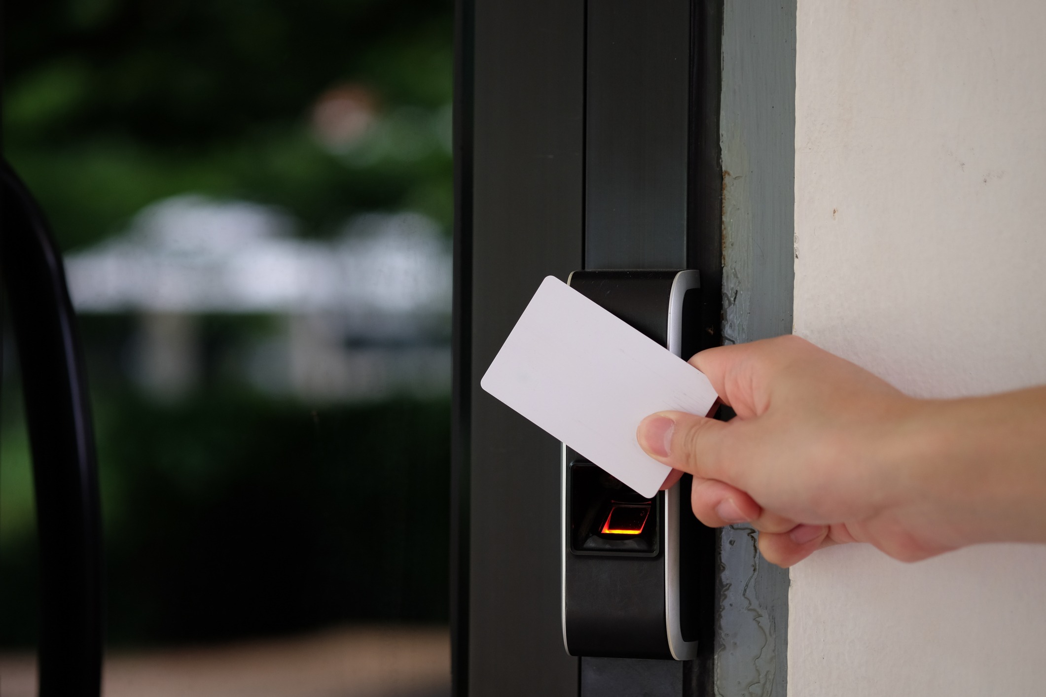 A person holds a white card near an electronic card reader, presumably for access. The background shows blurred greenery outside a building.