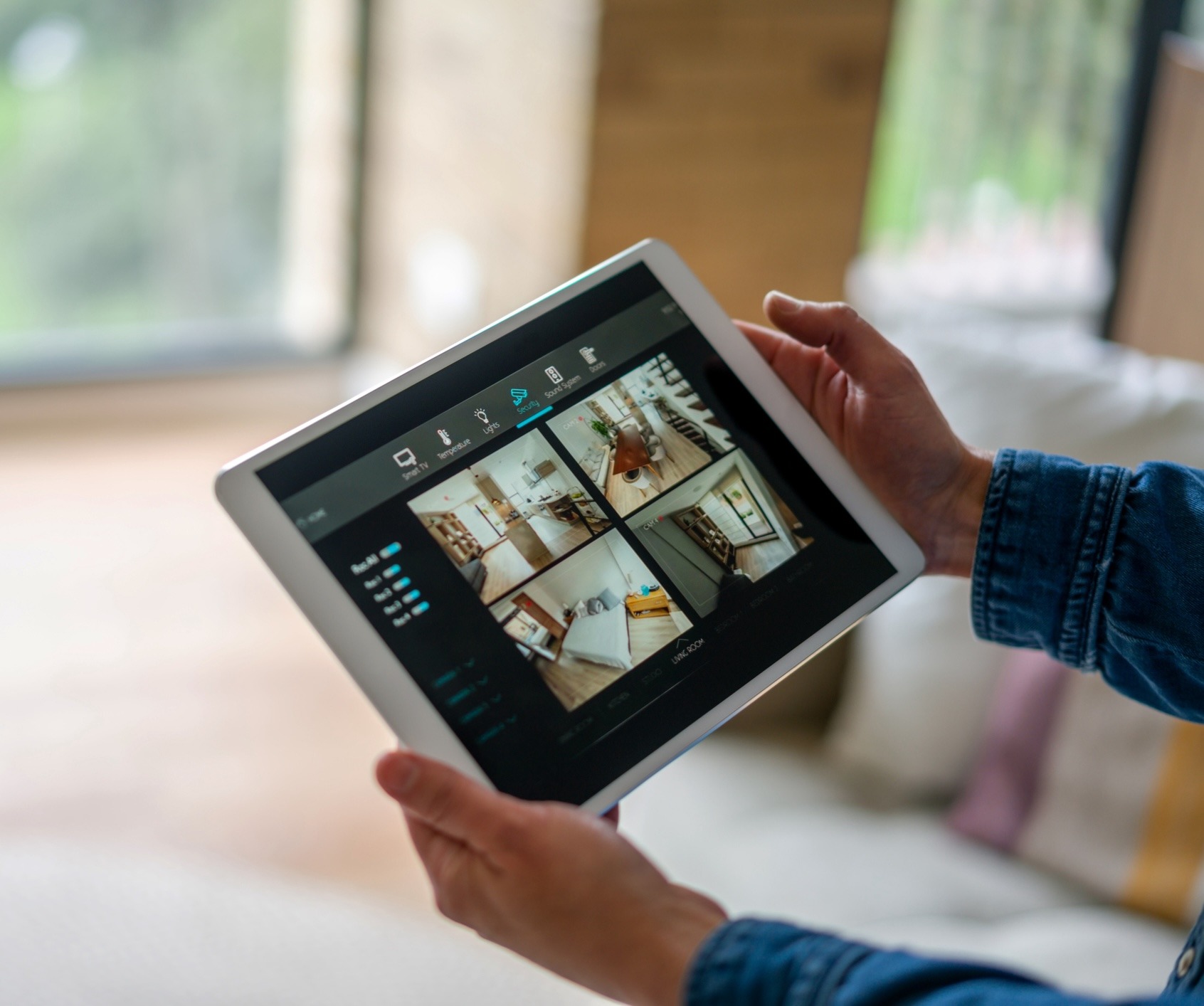 A person holds a tablet displaying a home security app with multiple camera views in a cozy indoor setting.