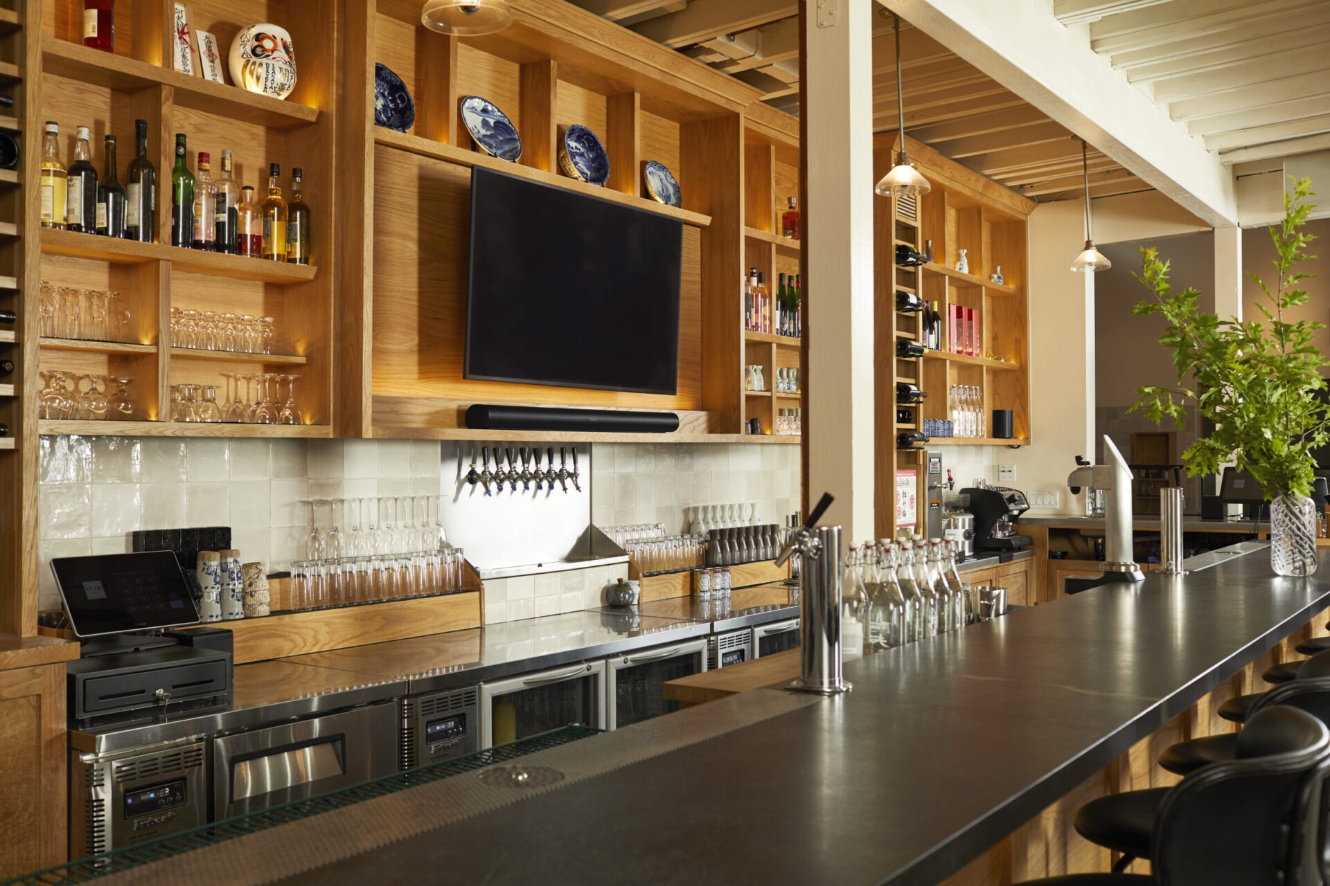 Spacious, modern bar with wooden shelves displaying liquor bottles and glassware. High ceiling, TV screen, and bar stools add to the inviting atmosphere.