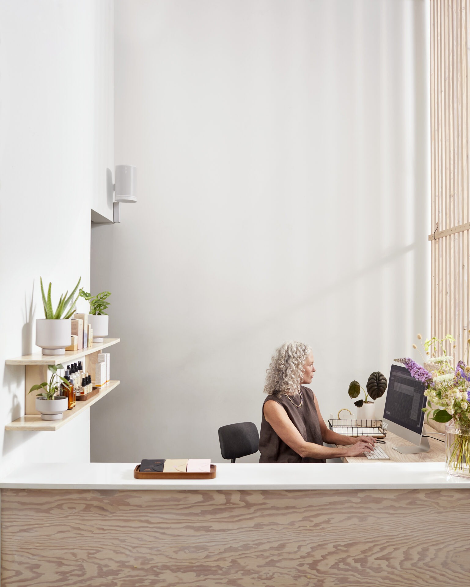 A person with curly hair works at a computer in a minimalist office, surrounded by plants and wooden shelves with small bottles.