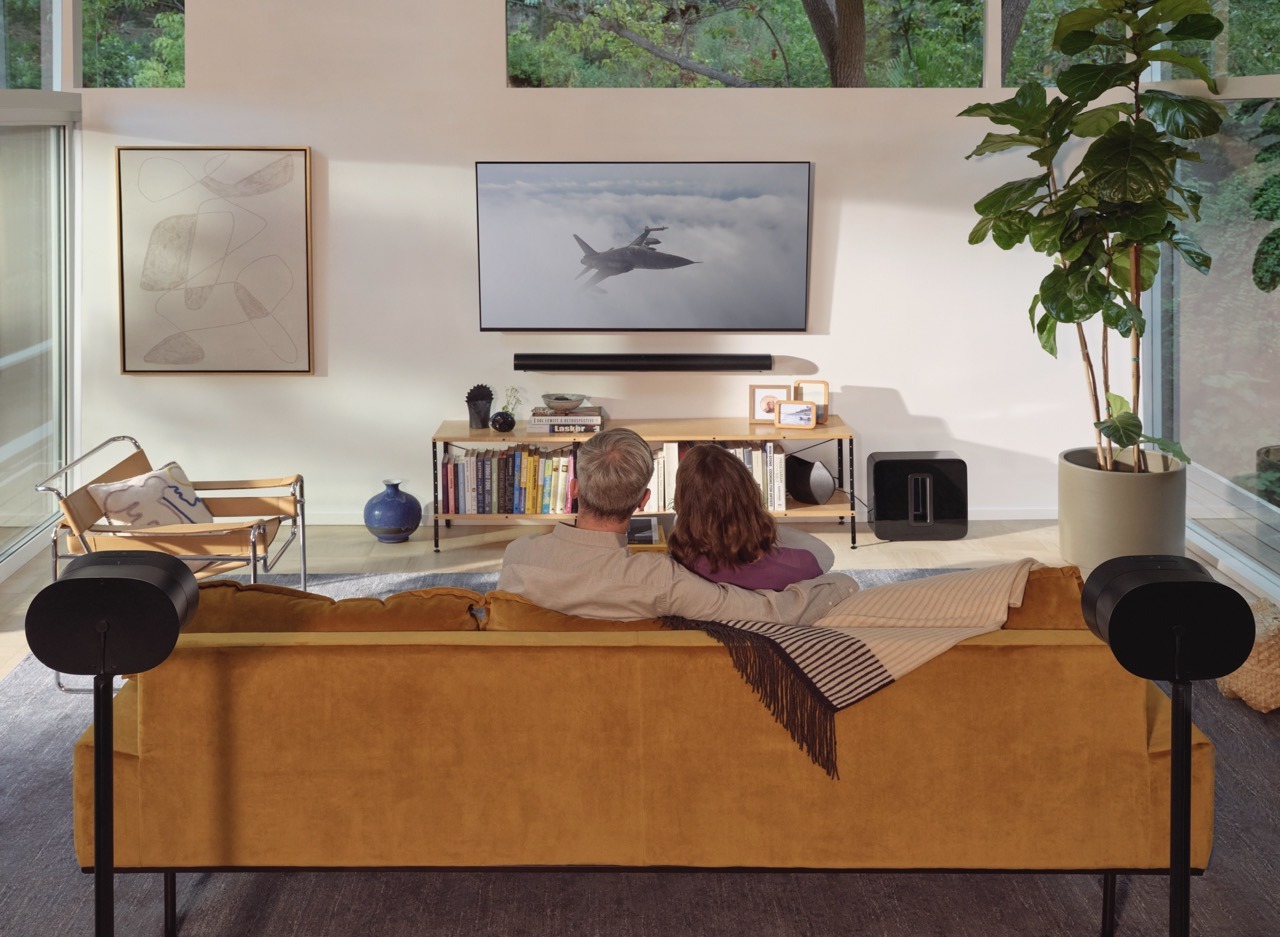 Two people are sitting on a yellow sofa, watching a jet on TV. The room has modern decor, including plants and wall art.