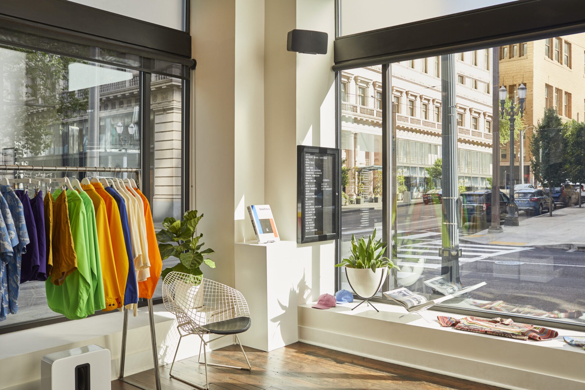 A modern retail store features colorful clothing and plants. Large windows showcase a city street with historic brick buildings outside.