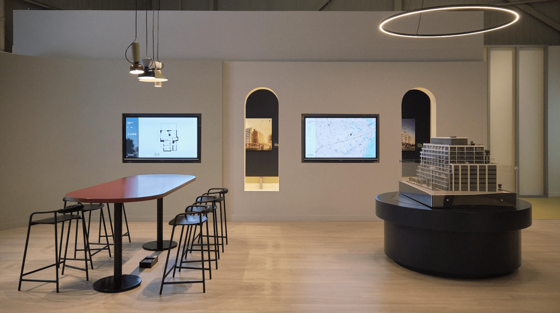 Modern exhibit room with architectural model, sleek red table, black stools, and digital displays on the wall, creating a minimalist design ambiance.