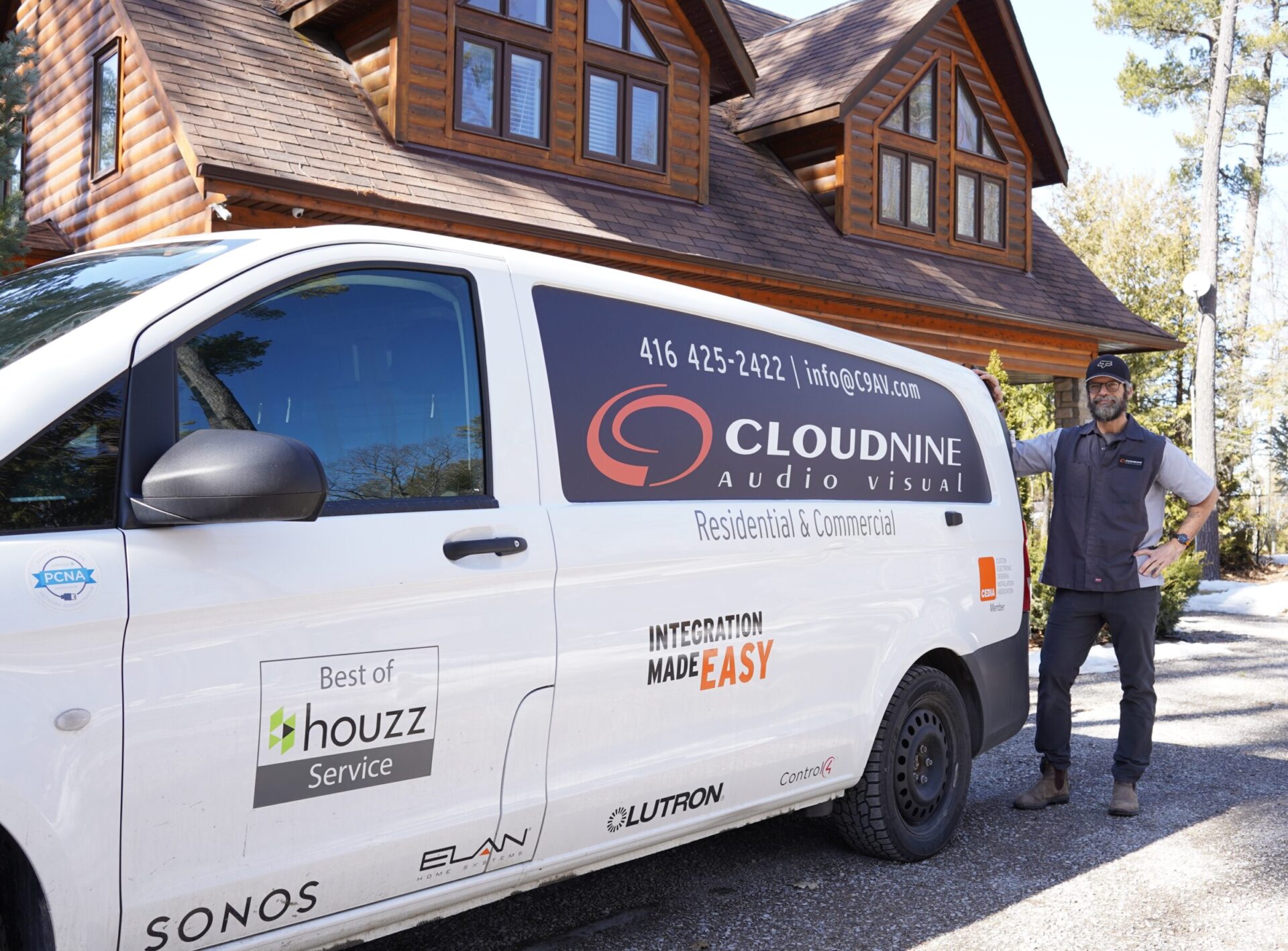 A person stands beside a Cloudnine Audio Visual van in front of a log house, surrounded by trees under a clear sky.