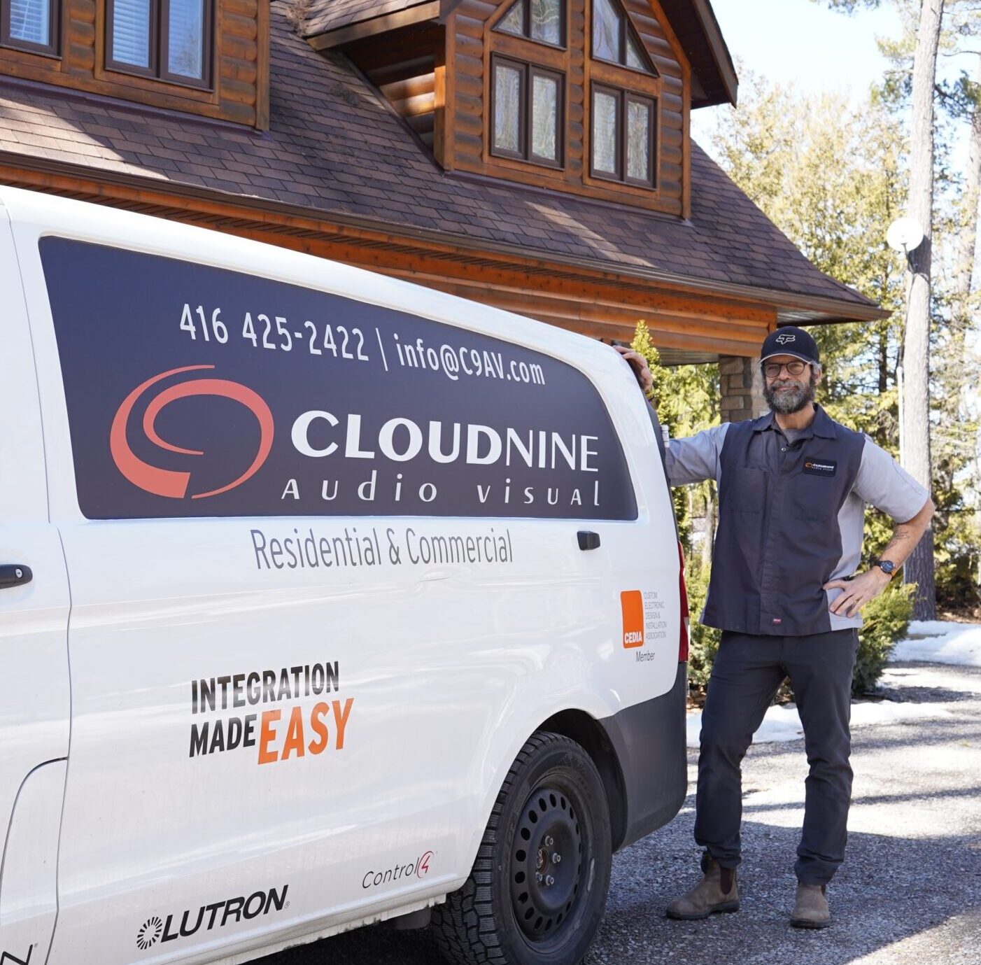 A person stands beside a Cloudnine Audio Visual van in front of a log house, surrounded by trees under a clear sky.