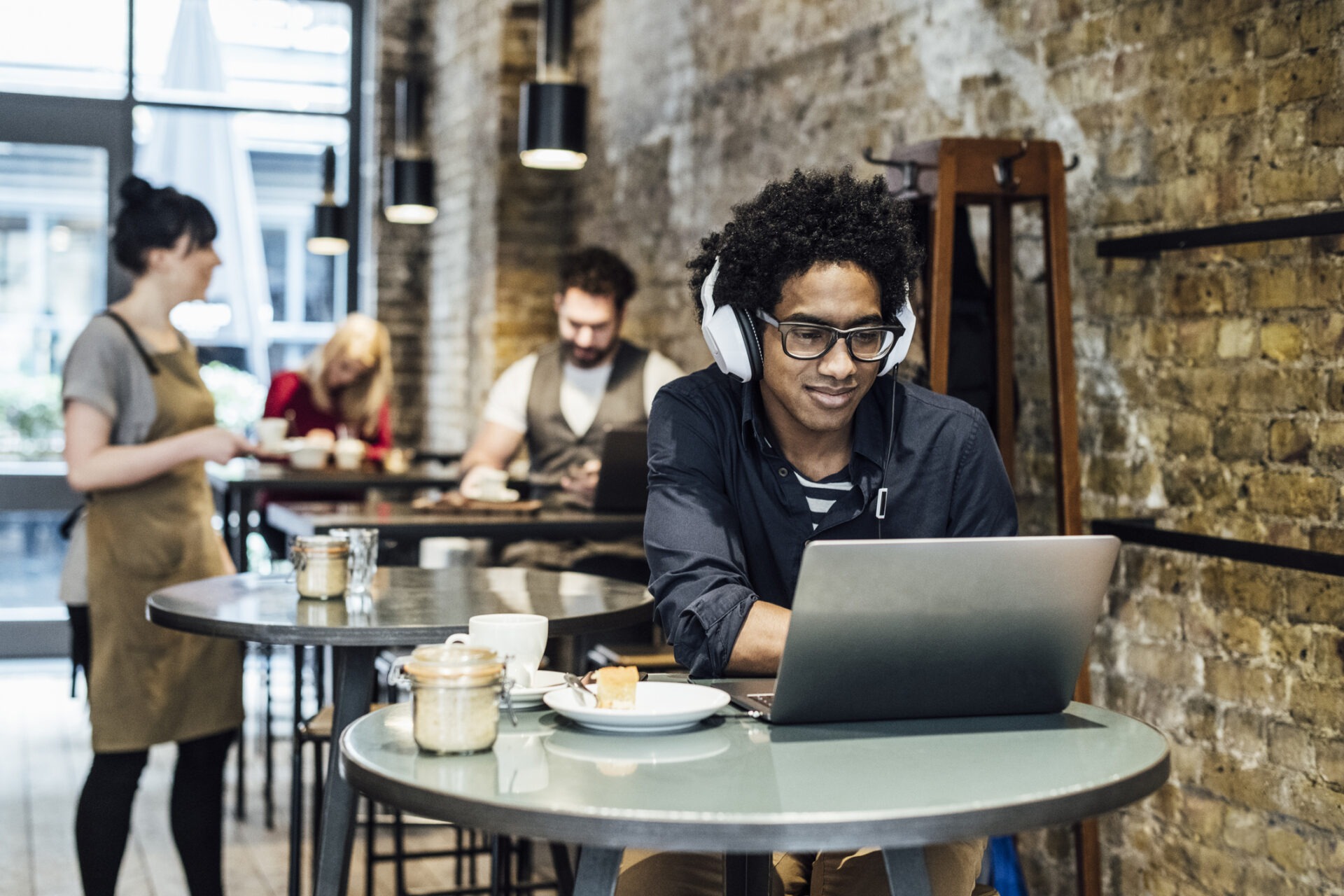 A person wearing headphones uses a laptop in a café. Other people in the background interact and work, creating a relaxed atmosphere.