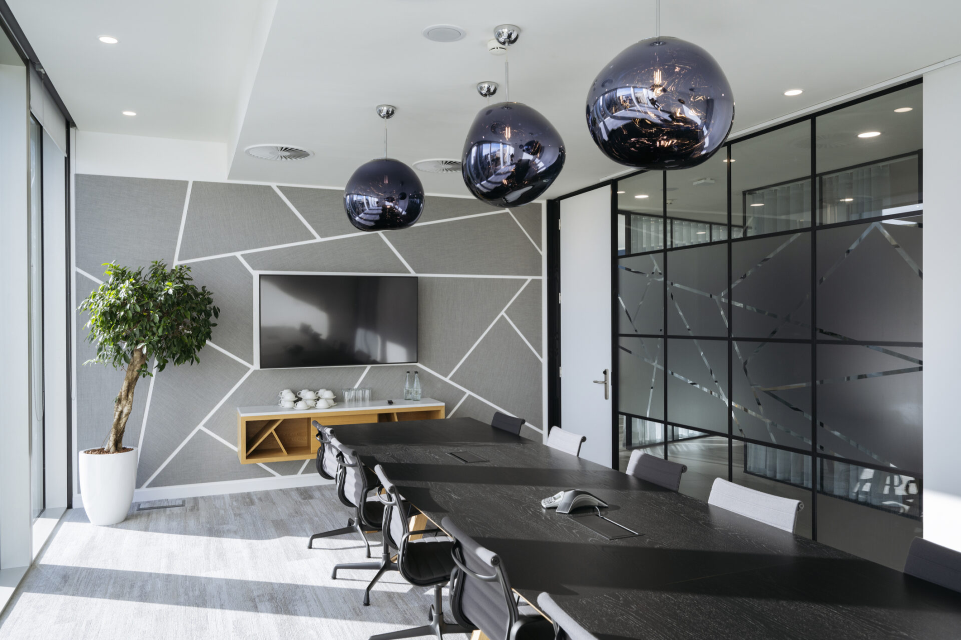 Modern conference room with large table, geometric wall design, and pendant lights. Glass partitions and potted plant enhance the contemporary office atmosphere.