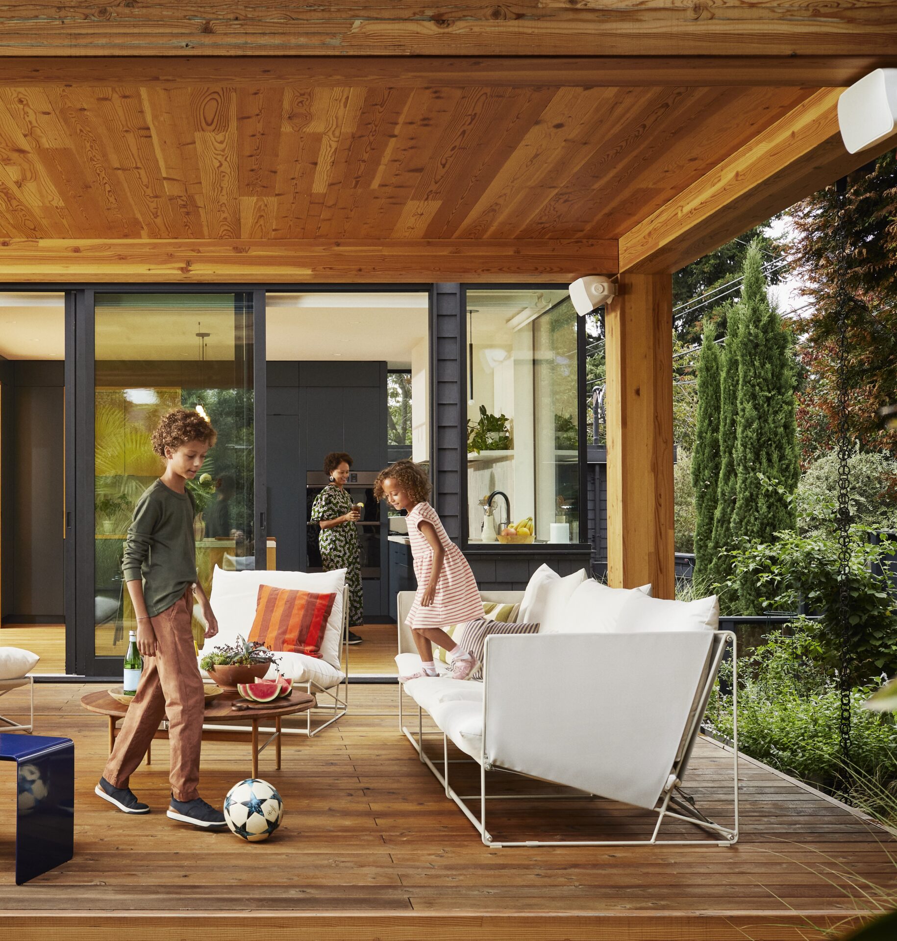 Modern outdoor patio with children playing soccer. A person is in the background inside the house. Wooden ceiling and glass doors enhance the scene.