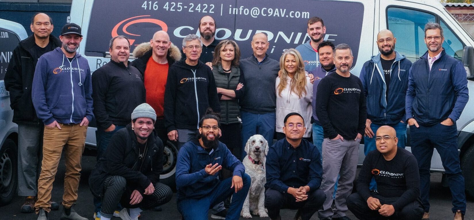 A group of people and a dog pose in front of branded vans for Cloud 9 AV, standing in an urban parking area.