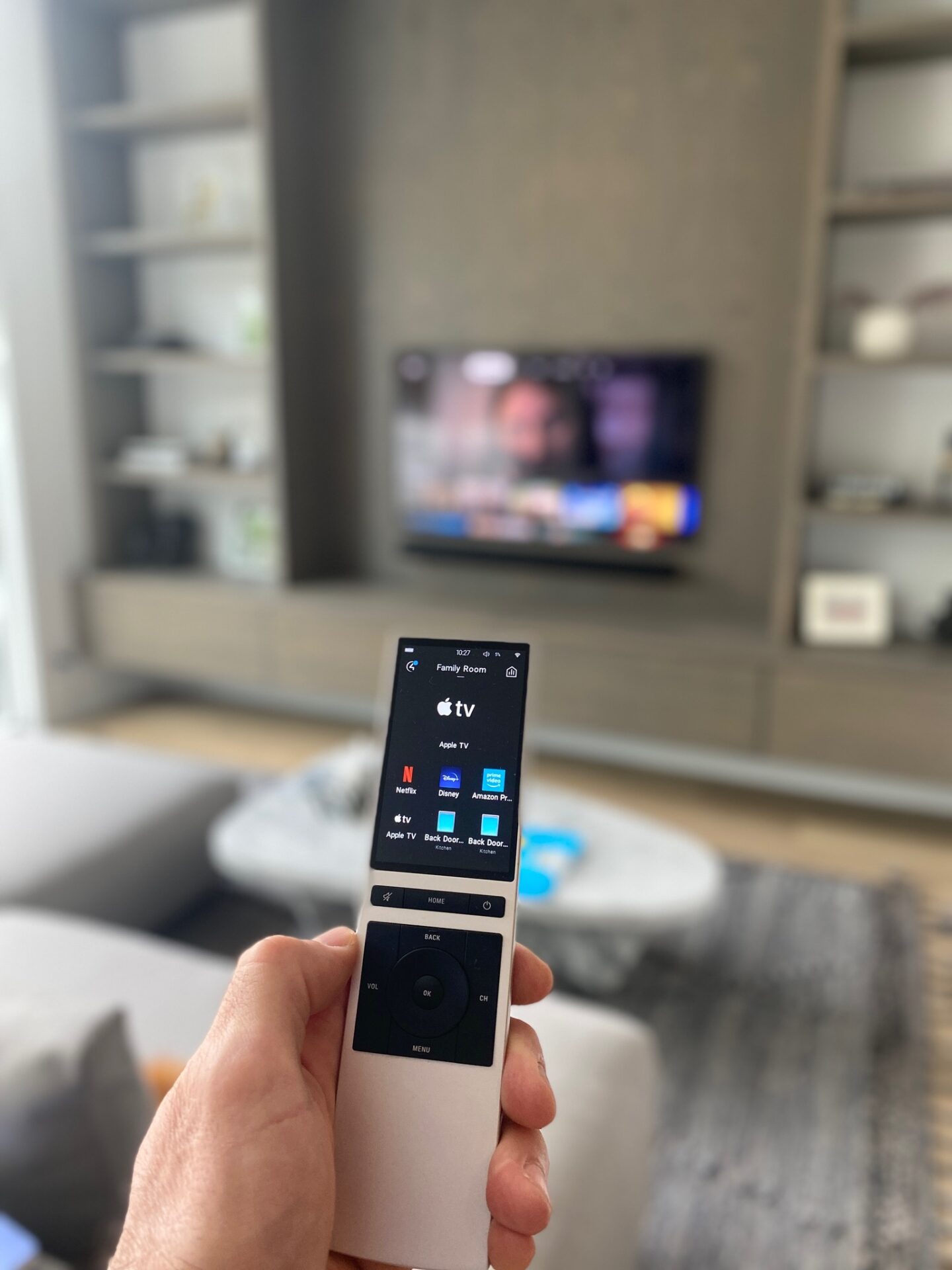 A person holds a remote control facing a wall-mounted TV in a modern living room with shelves and a gray color scheme.