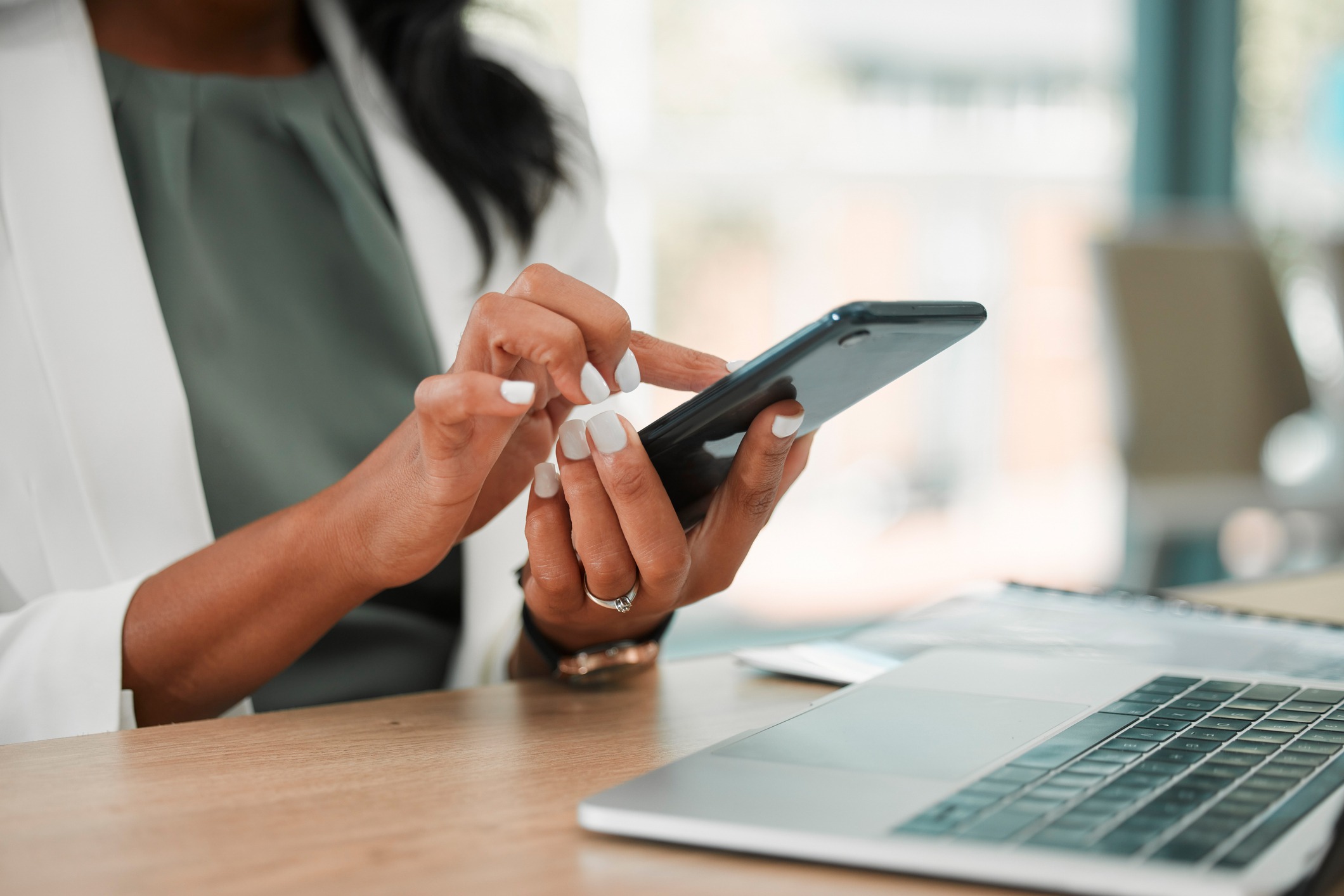 A person in a white jacket uses a smartphone at a desk with a laptop, in a modern office environment.