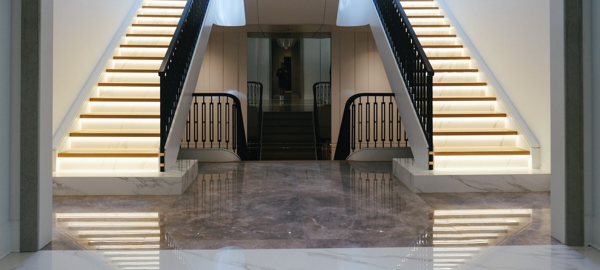 Elegant interior features dual illuminated staircases with black railings and marble floors. A person is visible in the background between the stairs.