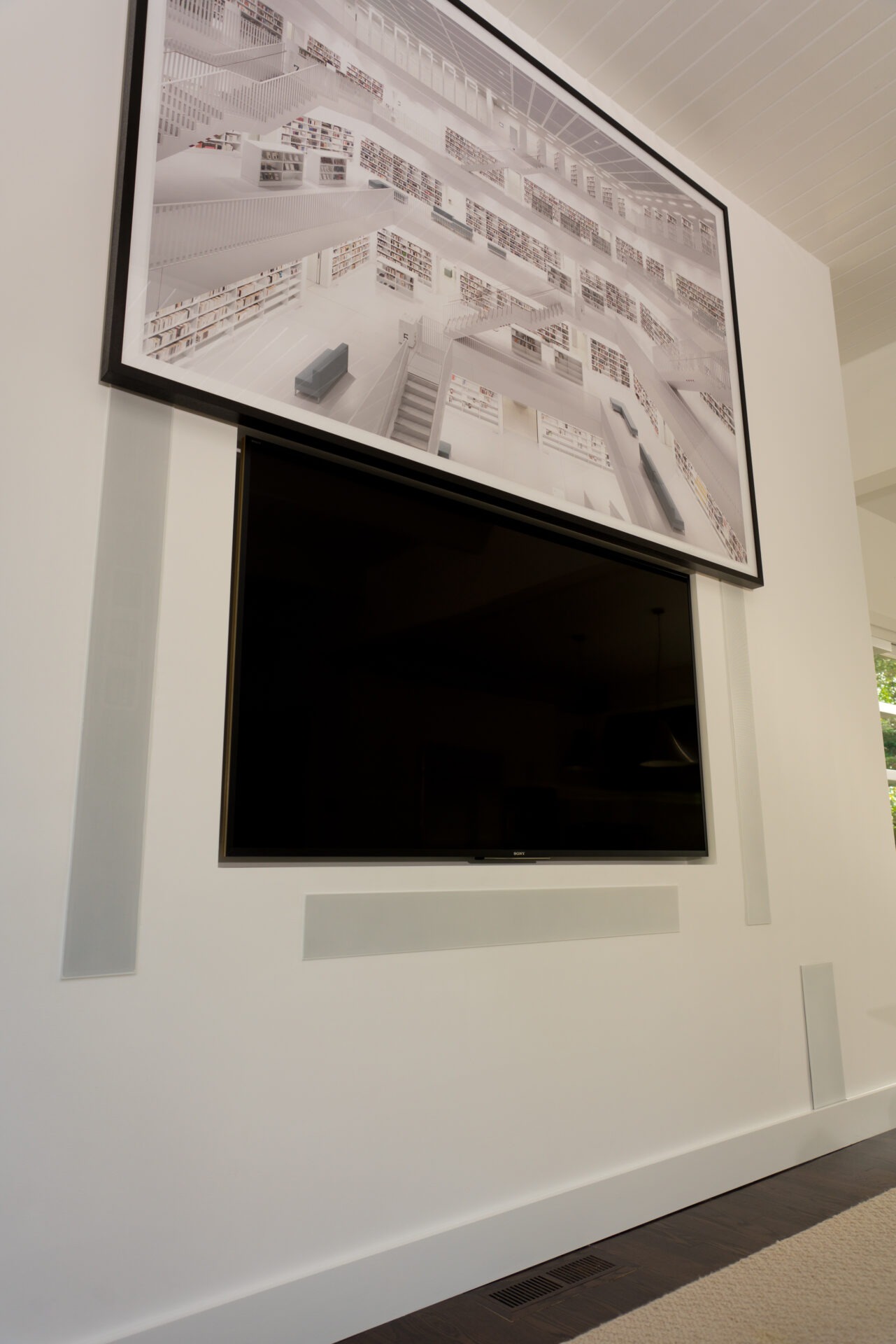 A large framed picture of a modern library is mounted above a flat-screen TV on a white wall with minimal decor.