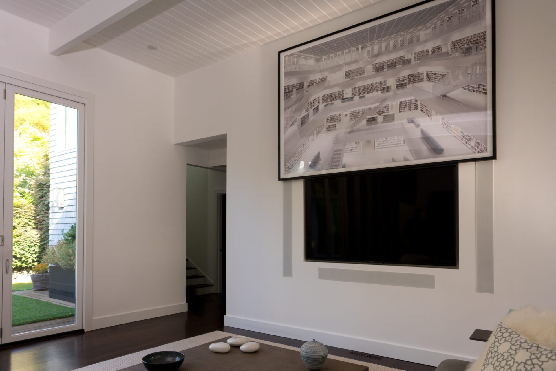 Modern living room with large wall art above a TV, featuring minimal furniture and garden view through sliding glass door. Bright, airy space.