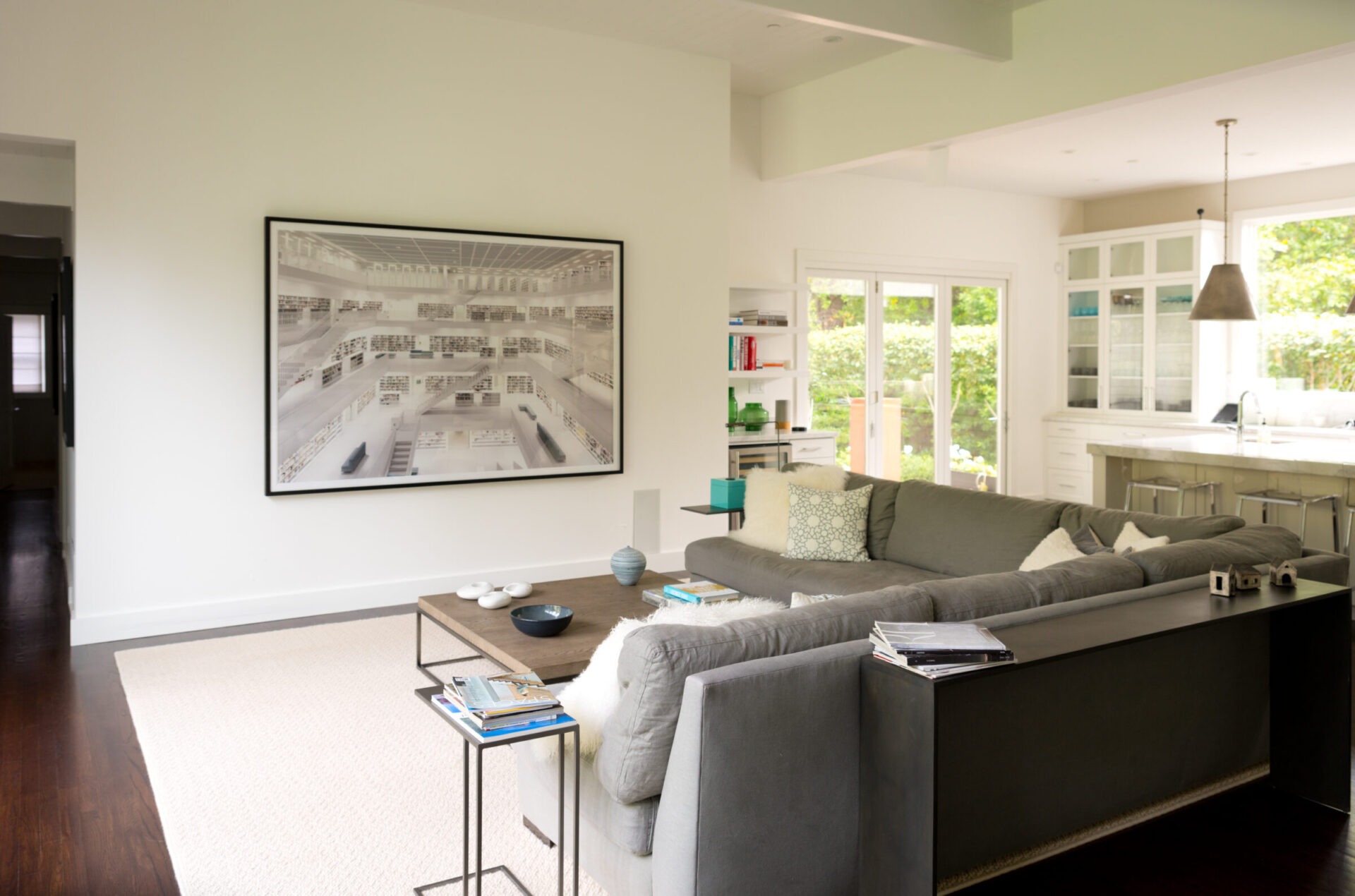 A modern living room with a large sofa, wall art, coffee table with books, and glass doors leading to a bright, leafy garden.