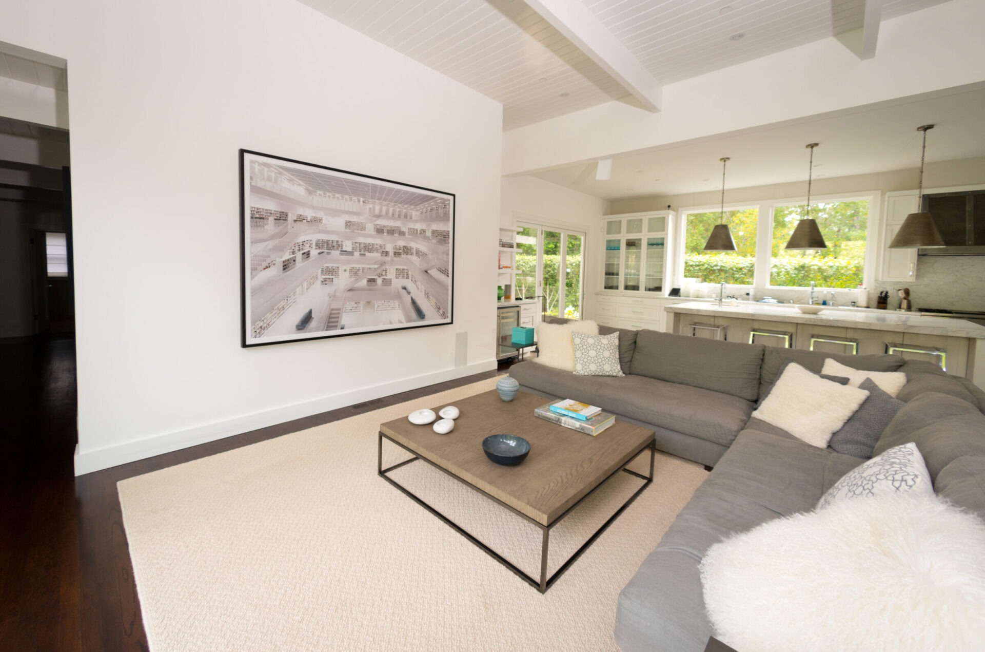 A modern living room features a large gray sectional, abstract artwork, minimalistic decor, and an open kitchen with pendant lighting and large windows.