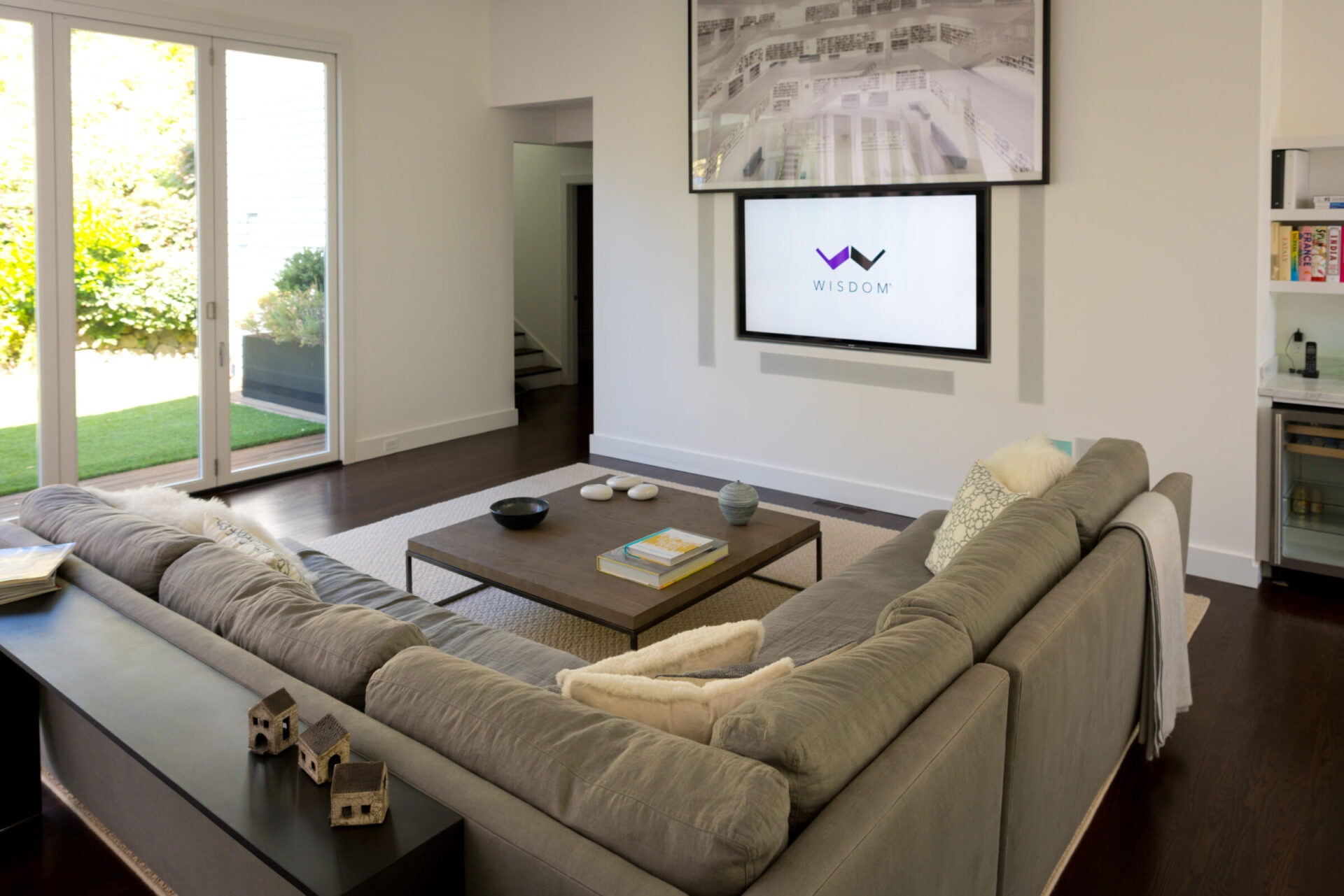 A modern living room with a large sectional sofa, coffee table, and two wall-mounted screens. Glass doors open to a garden.