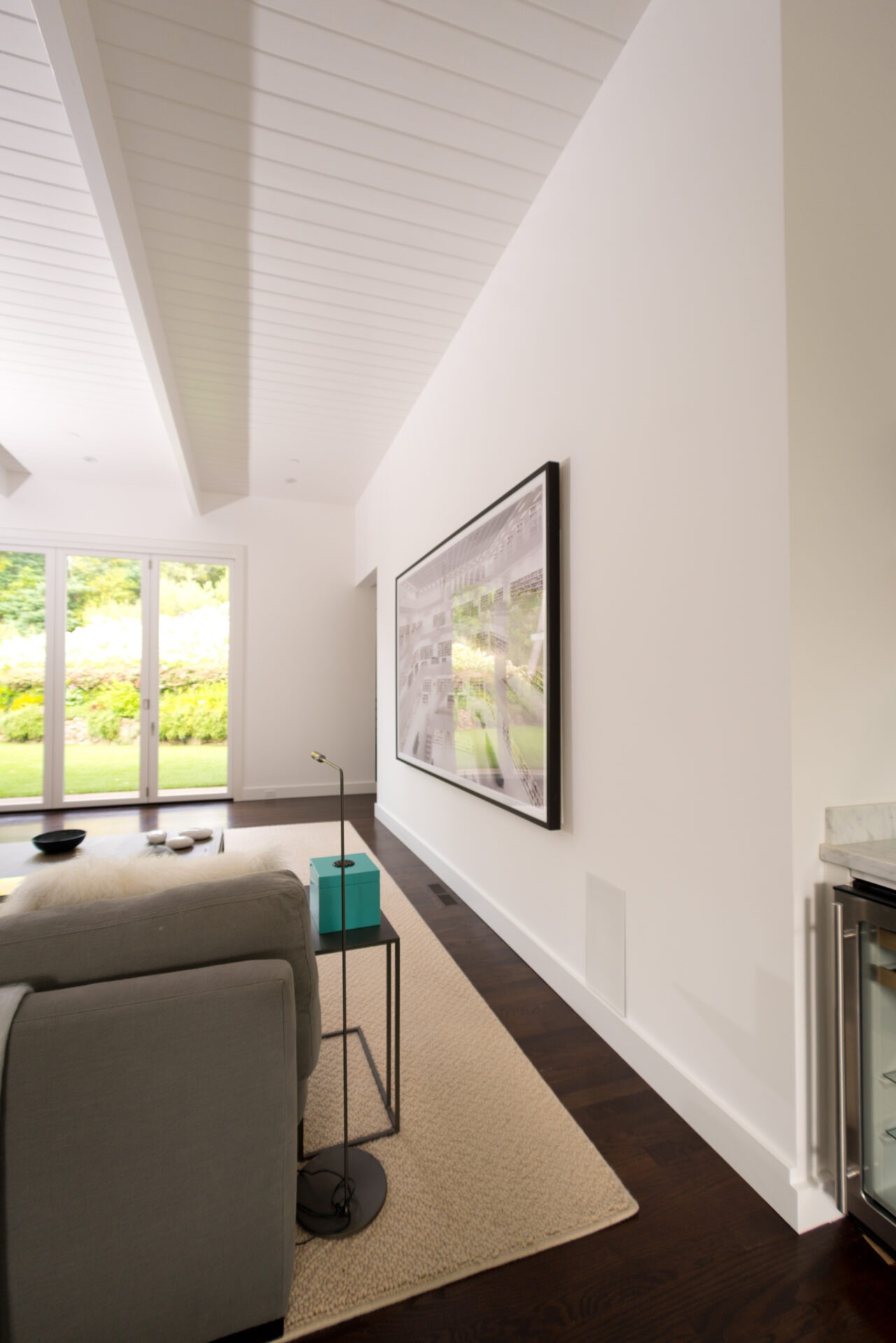 Modern living room with white walls, large framed artwork, glass doors leading outside, sofa, and side table. Natural light fills the room.