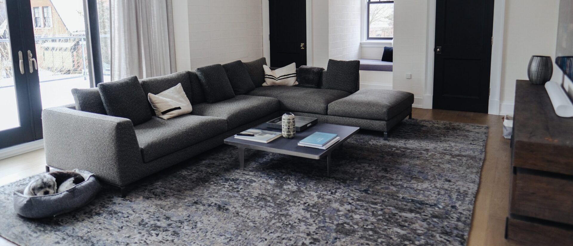 A modern living room features a large gray sectional sofa, coffee table, and large rug, with a cat sleeping in a corner.