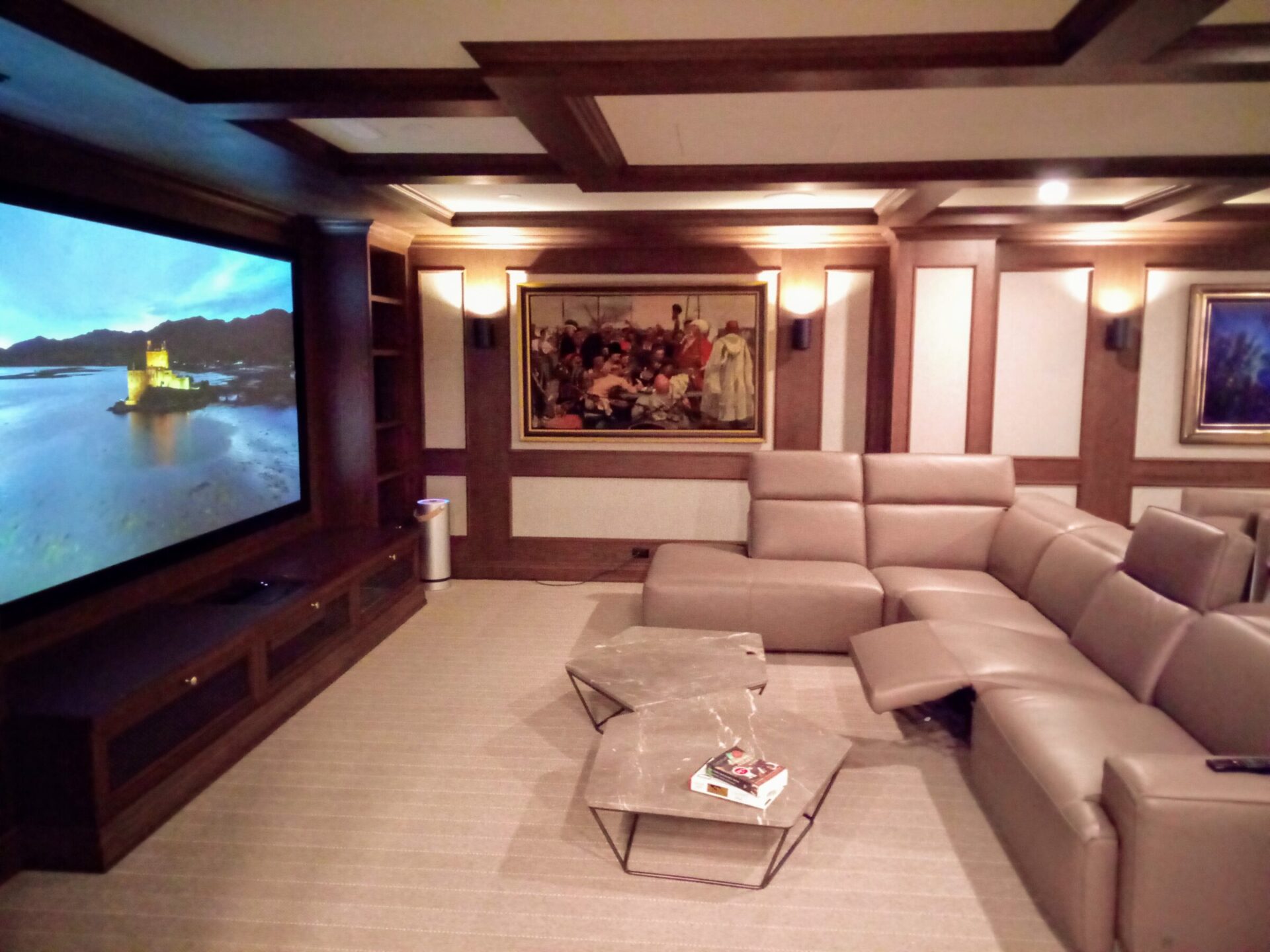 A cozy home theater features reclining seats, a large TV, shelving, and artwork. The screen displays a scenic waterfront view.