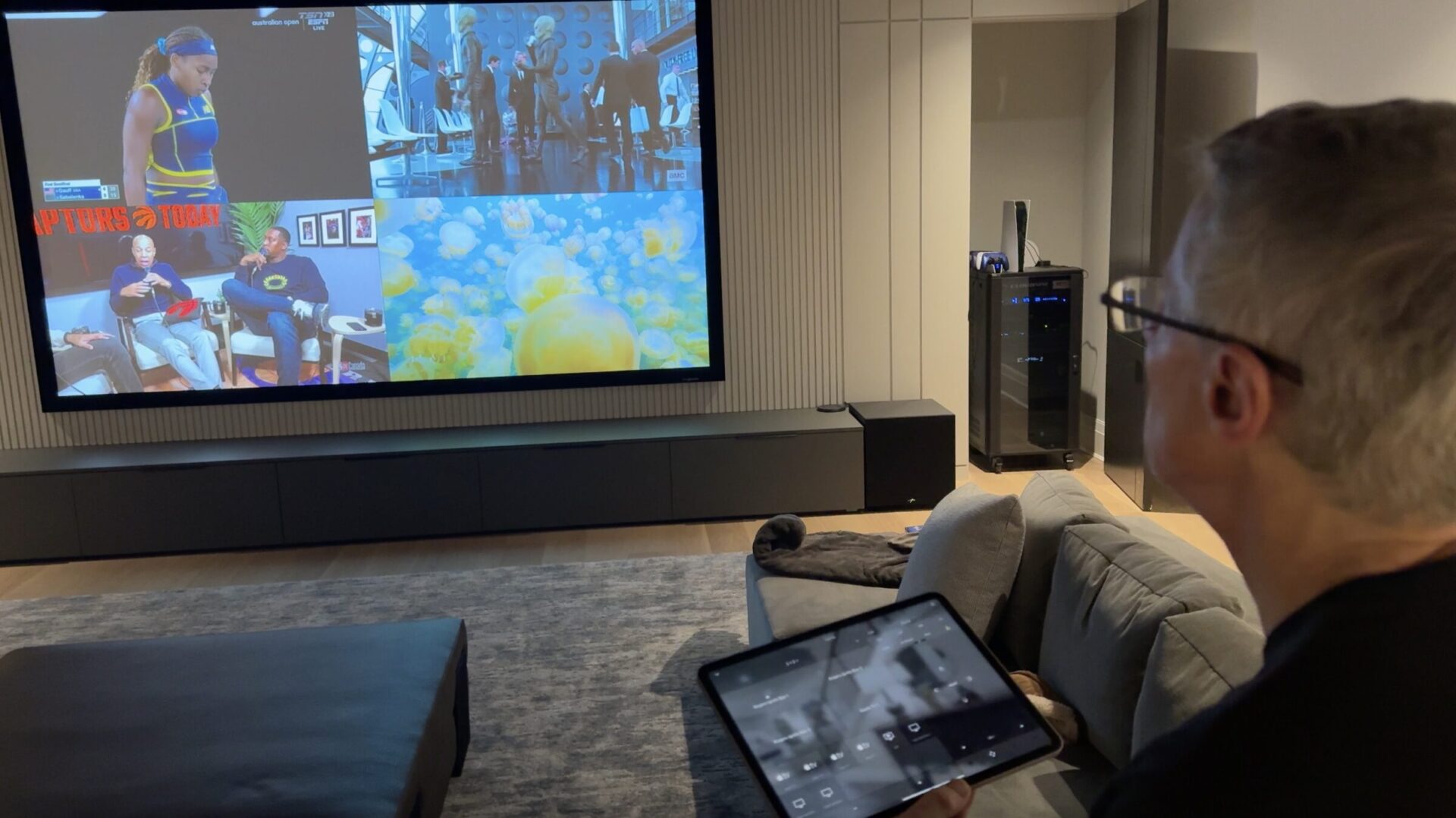 A person watching a large screen displaying four different channels, holding a tablet. The room is modern with minimalistic decor.