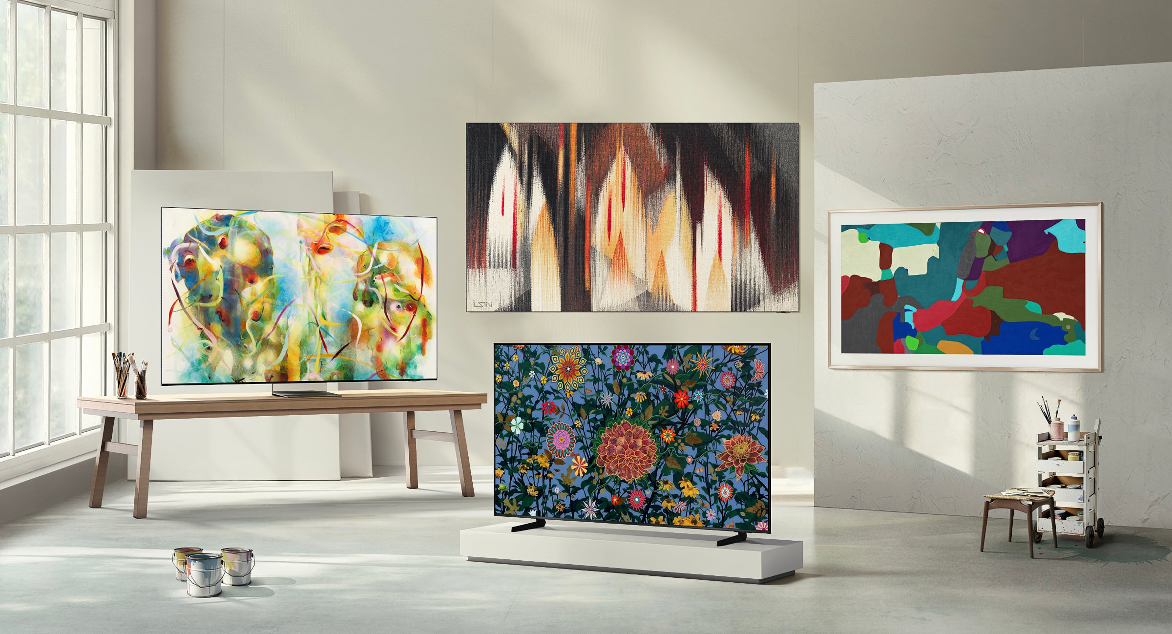 A room features four vibrant, abstract paintings displayed on two televisions and walls. Bright natural light from a large window illuminates the space.